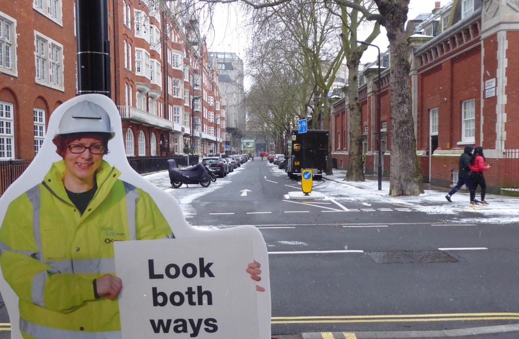 Looking down Bedford Avenue with a "cut-out" of a woman standing on the side of the road holding a "look both ways" sign.